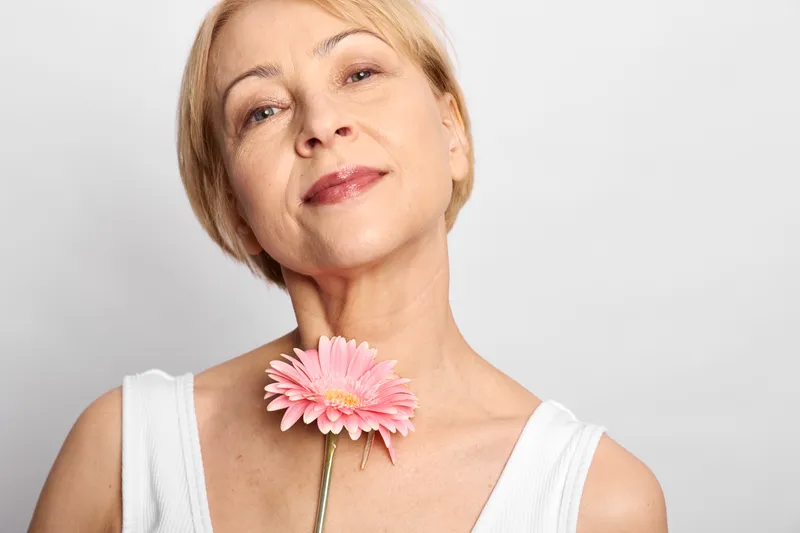 Mature woman closeup portrait smiling gently and holding a pink gerbera flower near her collarbone, studio isolated on white background, soft light