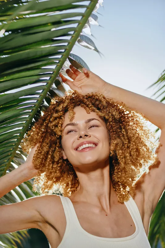smiling woman with curly hair enjoying sunny day near green palm leaves, bright natural light, relaxed mood, summer outdoor portrait