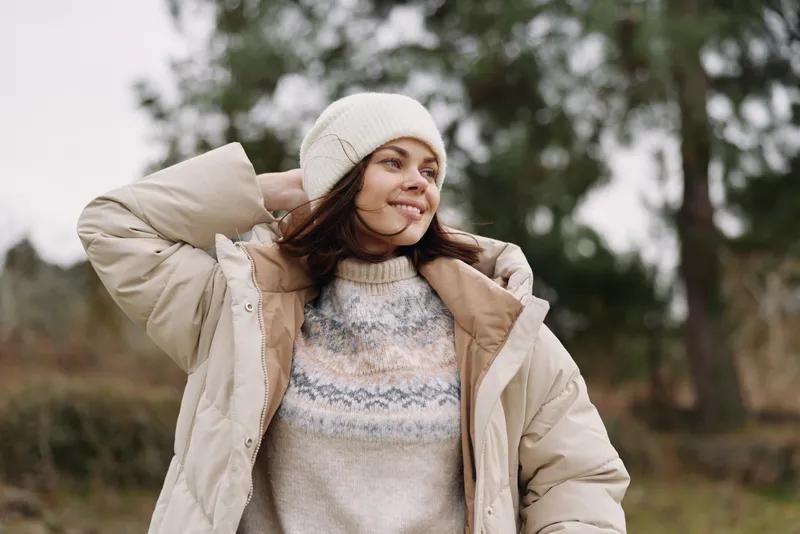 Woman in a cream jacket and beanie standing outdoors, cozy knit sweater visible, soft natural light, candid portrait capturing casual autumn style and relaxed mood.