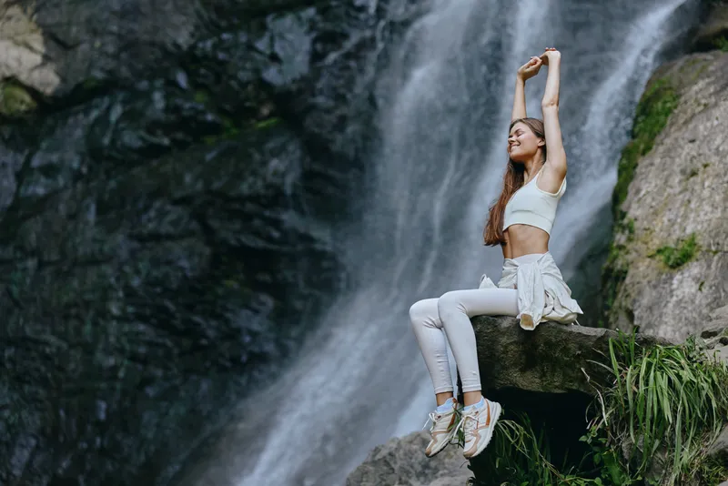 Woman stretching near waterfall, wearing white sportswear, sitting on rock, enjoying nature and fresh air in outdoor adventure setting.
