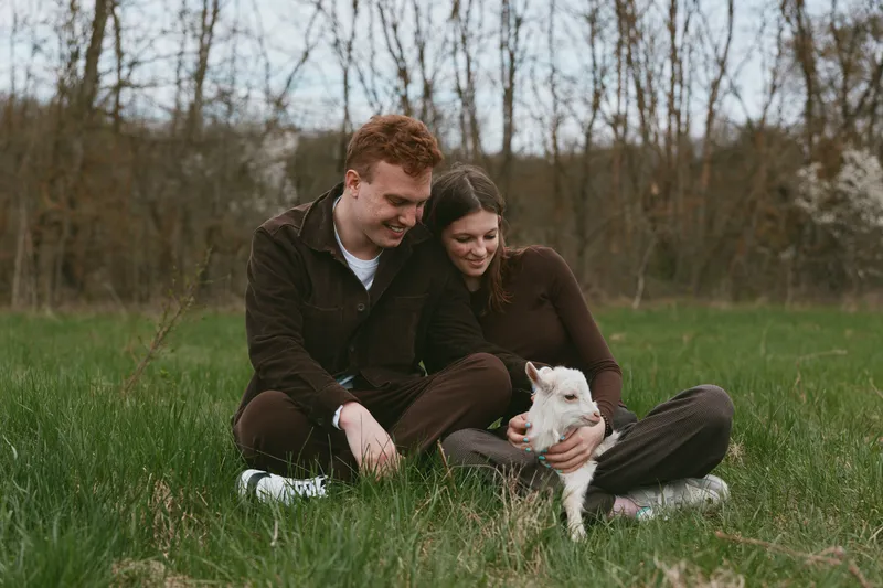 A couple sits on the grass outdoors, gently caring for a young goat kid, while a companion pet looks on. A lifestyle moment focused on warmth, connection, and rural calm.