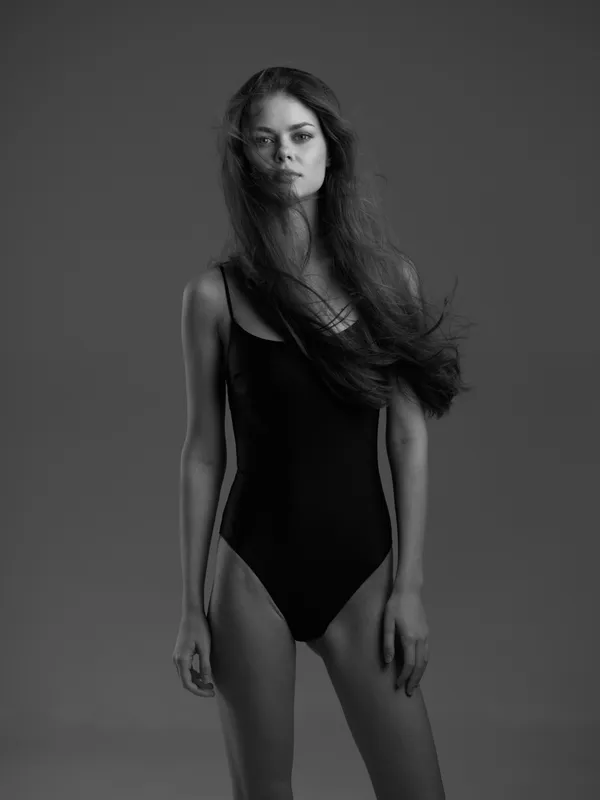 Black and white portrait of a woman with long hair in a black swimsuit standing confidently against a plain background.