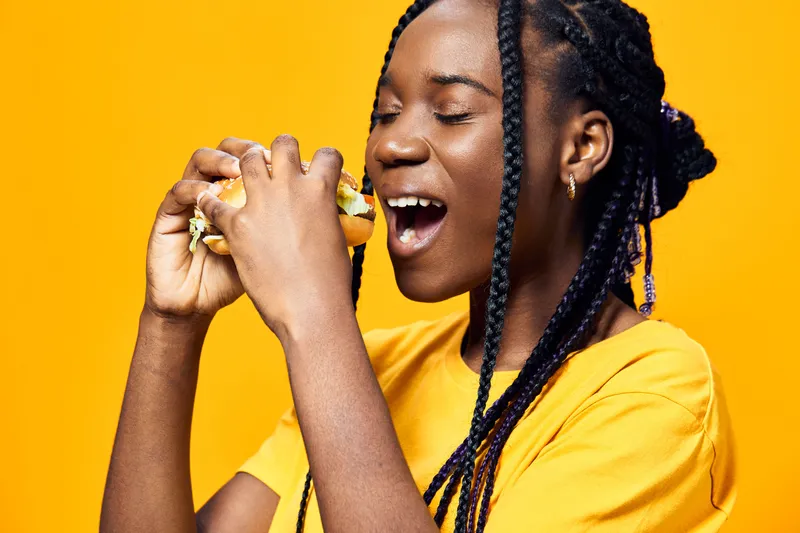 Young woman enjoying a delicious burger, cheerful expression, vibrant yellow background, showcasing healthy eating and happiness in a casual style