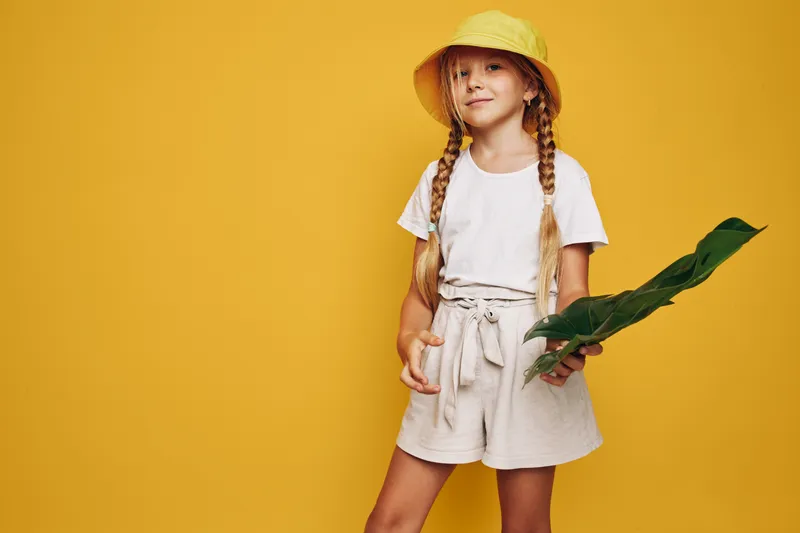 Young girl with long blonde hair in braids stands confidently wearing a yellow hat and white outfit holding large green leaves on a yellow background.