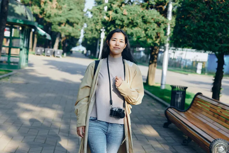 Woman in beige coat walking along a park path with a small camera hanging, casual street style, greenery, benches, day lighting, candid outdoor moment, fashion mood