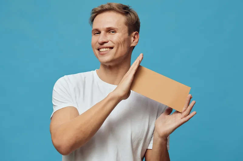 Smiling young man with casual white t-shirt holding a cardboard box against vibrant blue background, expressing happiness and surprise, studio lifestyle portrait.