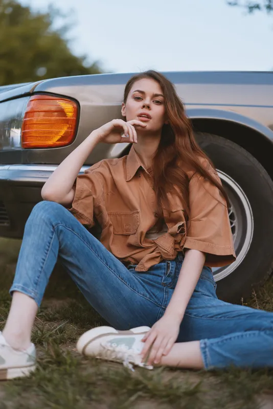 Young woman with long hair sitting beside a classic car, wearing a brown oversized shirt and jeans, exuding a relaxed and carefree vibe in a natural outdoor setting.