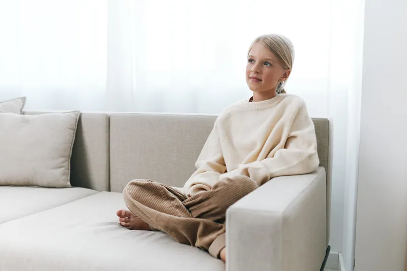 A young girl sits barefoot on a light sofa, wearing a soft beige sweater and brown pants, gazing upward with a calm, contemplative expression in bright room.