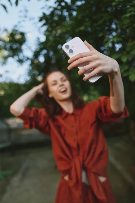 A cheerful woman in a red dress takes a selfie in a sunny outdoor setting, capturing a carefree lifestyle moment in a park with trees and blue sky