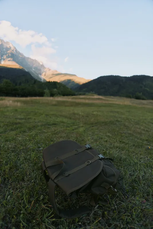 Meadow scene with a backpack resting on grass, distant mountains under a clear sky, calm outdoor lifestyle, natural colors, peaceful mood, landscape focus, hint of adventure