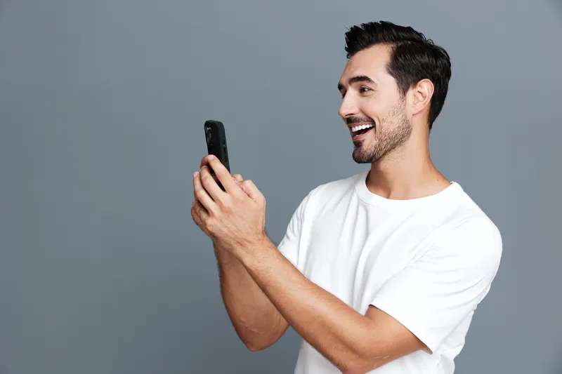 Smiling man using mobile phone while looking at screen, enjoying moment and capturing memories, dressed casually in a white t-shirt, set against a gray background.