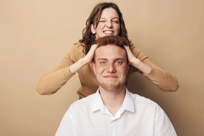 Playful young woman in beige sweater squeezes the head of smiling redhead man in white shirt. Studio portrait on beige background. Concept of friendship, fun, and casual lifestyle.