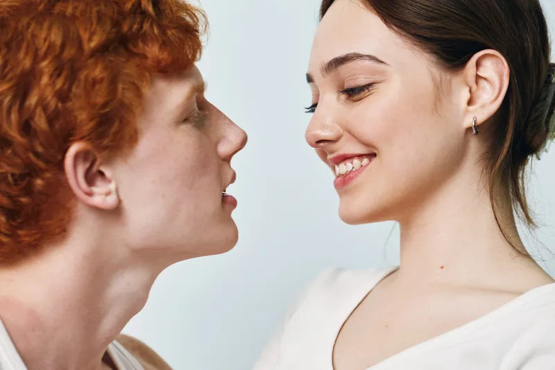 Young man and woman facing each other, smiling and about to kiss with closed eyes and happy expression. Intimate moment, closeup portrait with light background and natural skin tones.