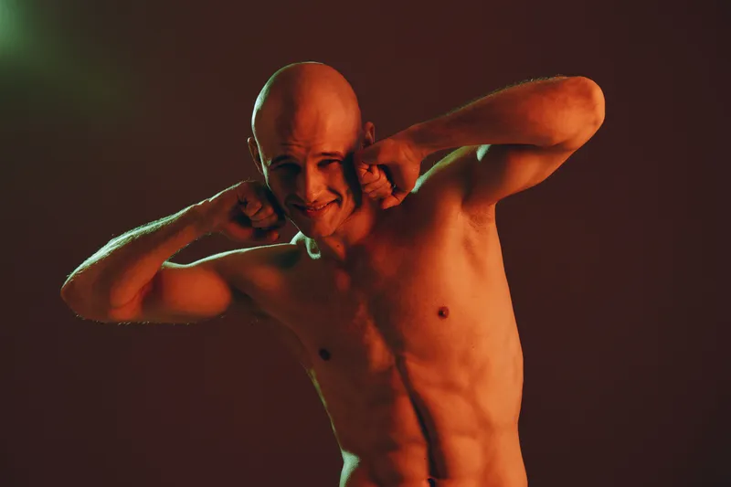 A muscular man with a bald head playfully covering his ears, expressing fun and excitement in a dynamic pose against a warm, colorful backdrop.