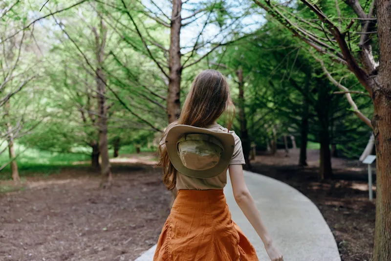 Woman walking alone in park among tall trees on a winding path, wearing a hat and orange skirt, back view of solo traveler enjoying nature and peaceful green woodland.