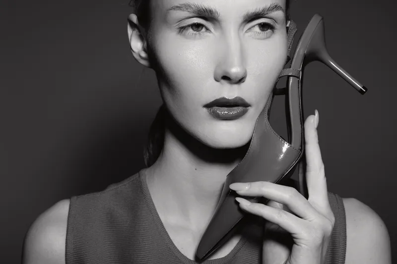 Caucasian woman with pale skin holds high heel shoe near face with serious expression in black and white photo.