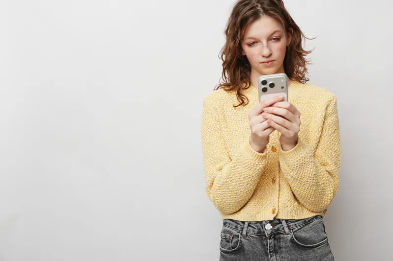 Young woman in yellow sweater looks at smartphone with focused expression. Casual style, isolated on white background. Concept of communication, technology, and social media use.