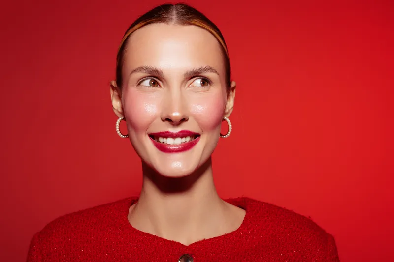 Smiling woman with red lipstick and hoop earrings looks to the side on red background. She wears a red sweater, creating a vibrant and confident portrait with bright studio lighting.