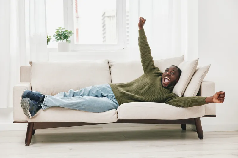 A joyful Black man relaxing on a cozy sofa, celebrating life with wide smiles and open arms, in a bright and minimalist living room