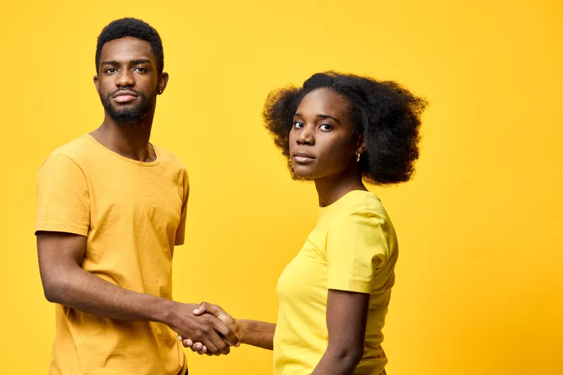 Two young adults shaking hands against a vibrant yellow background, dressed in matching yellow shirts, expressing confidence and professionalism
