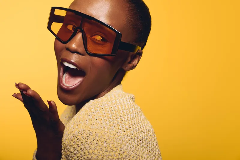 Happy African woman with dark skin wearing large sunglasses and yellow sweater poses on yellow background. Concept for fashion, joy, style, and confident female model portrait.