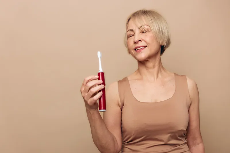 Smiling mature woman with short blonde hair holds red electric toothbrush against beige background. Concept of dental hygiene, oral care, healthy lifestyle, and self-care for senior adults.