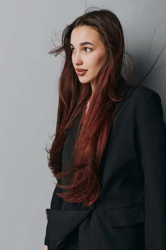 Young woman with long red hair wearing black blazer standing against gray wall looking to side with natural makeup and loose hair for fashion portrait and beauty concept.