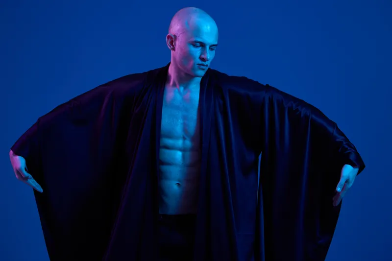 Striking man in dramatic pose, showcasing a toned physique, dressed in flowing black robe, set against a captivating blue background.