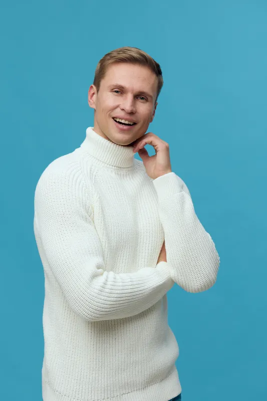 Smiling young man with blond hair, wearing a white turtleneck sweater, stands confidently against a bright blue background. Happy expression, casual style, cheerful mood, portrait photography, studio 