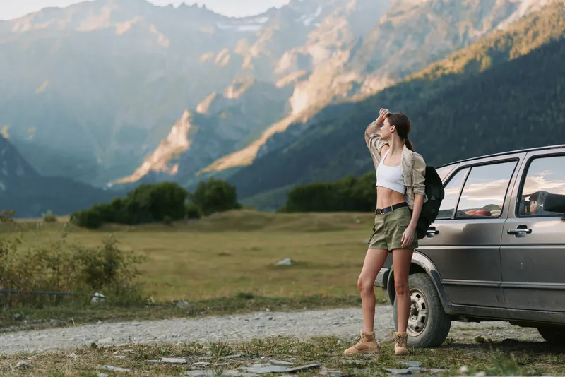 Woman traveler beside a car in a mountain landscape, stretching after a hike. Casual outfit, backpack, open road, fresh air. Outdoor exploration and wellness vibe.