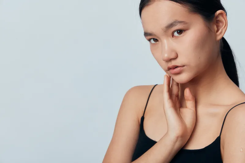 Young woman with long dark hair and natural makeup, posing with hand on neck in black sleeveless top against light gray background studio. Beauty, calm expression and skin care portrait.