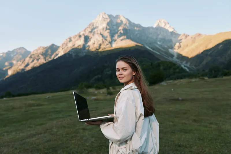 A joyful woman stands outdoors with a laptop in hand, surrounded by rugged mountains. She faces the camera with a calm, confident smile, projecting focus, creativity, and professional momentum in