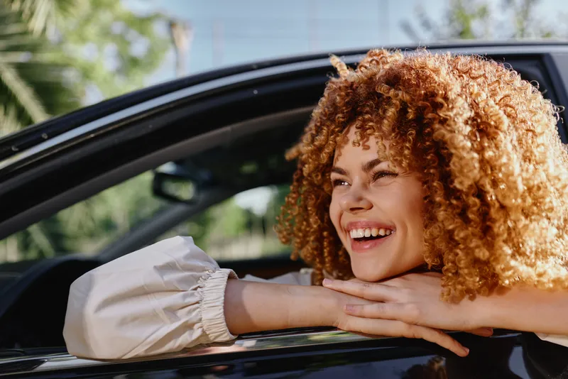 smiling woman with curly hair leaning out car window, enjoying sunny day with natural light and green background, expressing happiness and relaxation