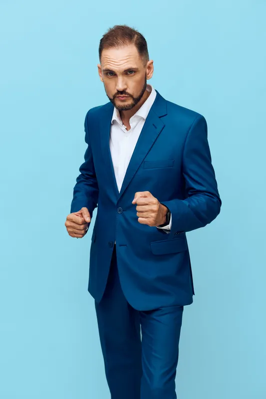 A professional business man in a blue suit stands against a solid colored background, projecting confidence, determination, and readiness.