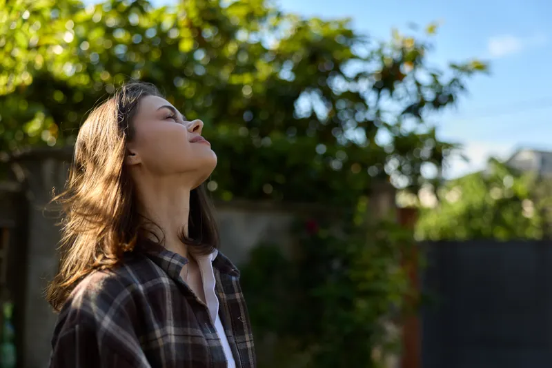 Smiling woman with shoulder length hair standing outdoors, head tilted toward the sun. Casual plaid shirt, nature backdrop, feeling relaxed, carefree, and reflective