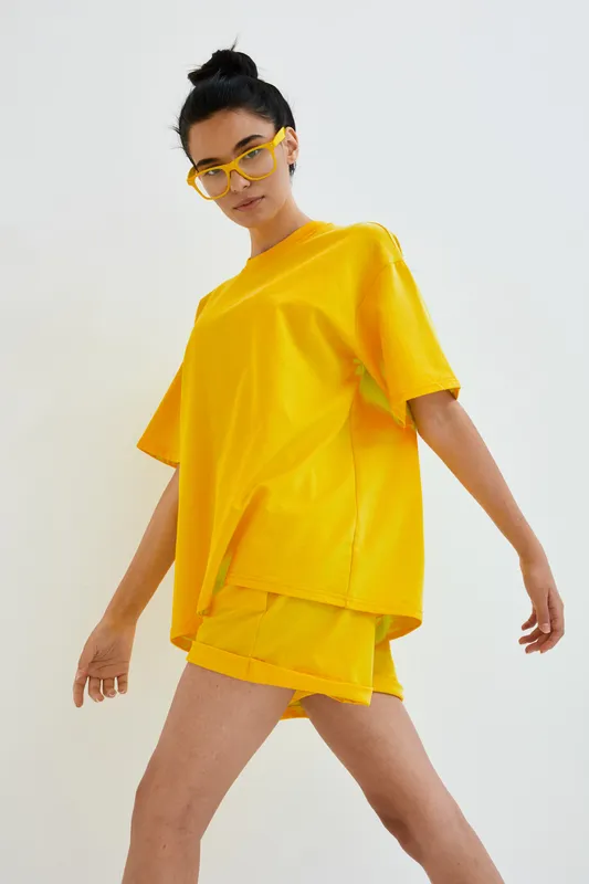 Young woman wearing bright yellow oversized t-shirt and shorts set with stylish sunglasses posing confidently against white background, minimalistic fashion look.