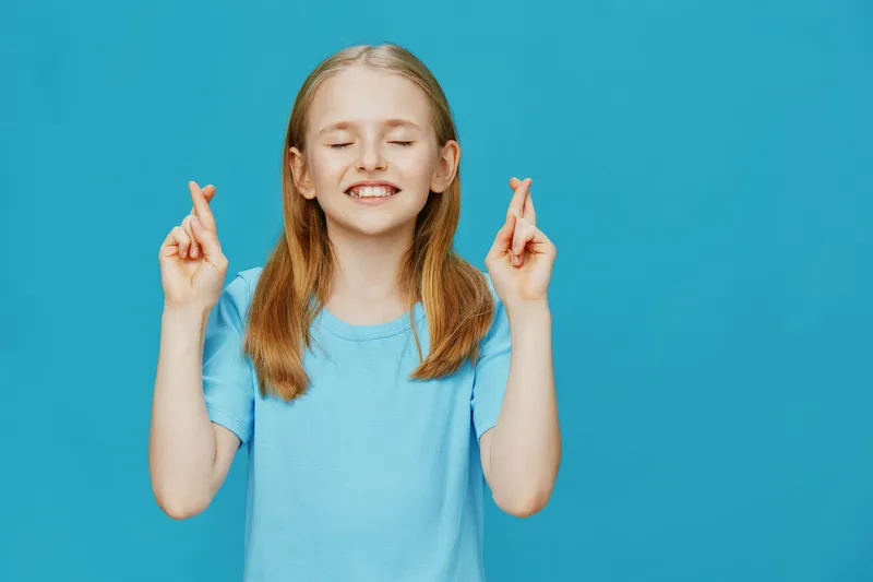 Cheerful young girl with crossed fingers, expressing hope and excitement against a turquoise background, wearing a light blue T shirt, ideal for positive concepts
