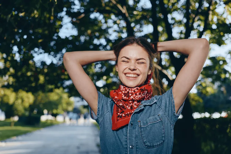 Lifestyle woman in modern boho-western denim with red bandana, smiling outdoors in street style, capturing film color warmth and casual happiness under green trees.
