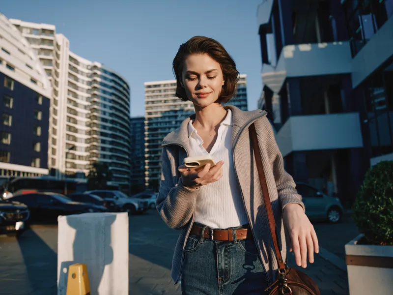 A stylish woman walks outdoors in an urban setting, smiling as she checks her phone. She wears a white shirt, cardigan, jeans, and a brown belt, projecting a modern, confident daily routine.