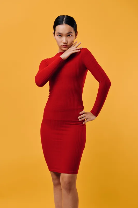 Young woman in a red fitted dress posing with hand near face against yellow background. Fashion model with sleek hairstyle and confident expression in studio lighting.