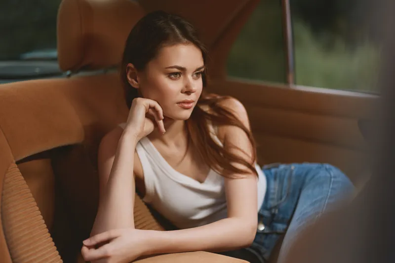 Young woman lounging in a tan car interior, wearing a white tank top and denim shorts, exuding a relaxed and contemplative vibe amidst a natural backdrop.