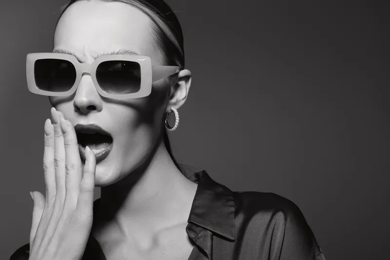 Stylish white woman with pale skin wearing large sunglasses and silky shirt yawns with hand near mouth in black and white studio portrait.
