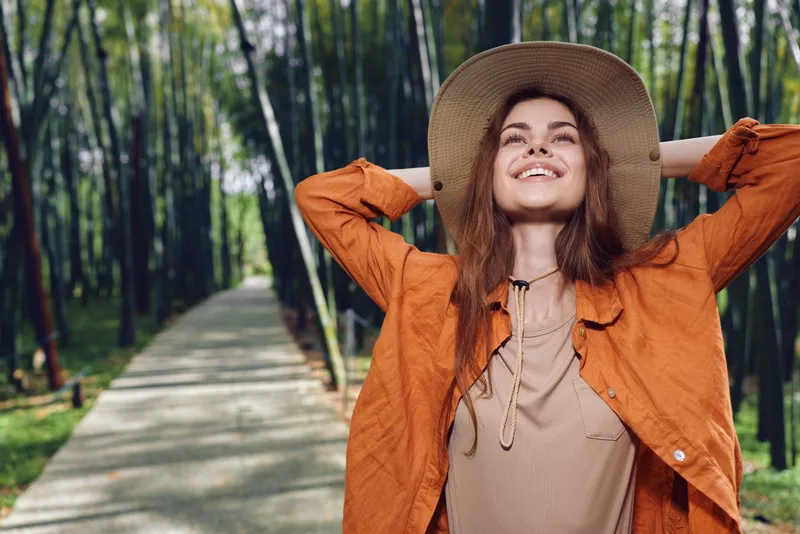 Woman with hat and smile in a forest path, portrait of a happy traveler relaxing with hands behind head. Nature walk, outdoor lifestyle in orange jacket among green bamboo trees.