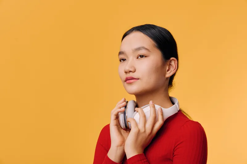 Young woman with headphones holding them near face, standing against orange background. Casual portrait of female with dark hair in red long sleeve shirt looking aside thoughtfully.