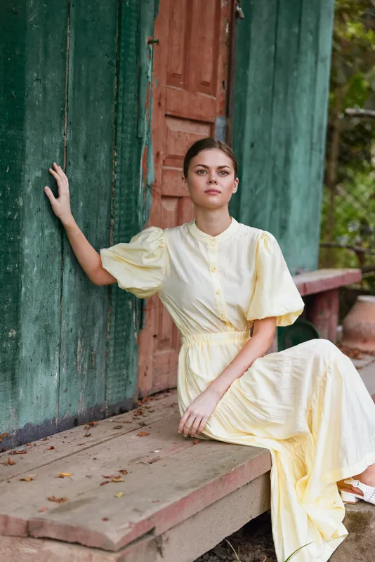 Woman in a pale yellow dress sits on rustic steps beside a weathered door, calm pose in an outdoor setting, soft natural light, tranquil and graceful atmosphere.