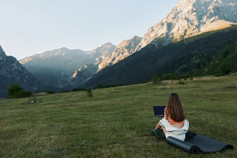 A focused traveler sits on a mat in a tranquil mountain meadow with a laptop, embracing remote work, nature, and quiet productivity in a scenic outdoor setting.