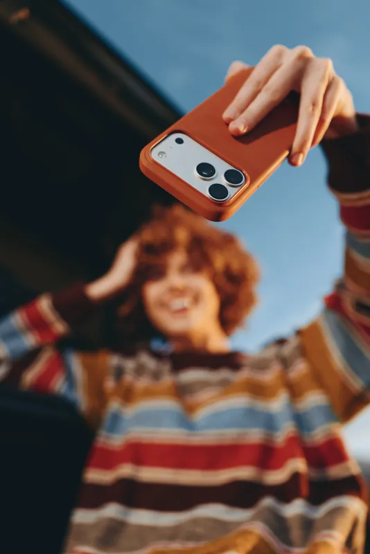 Woman smartphone lifestyle outdoors, hand holding orange phone in foreground, smiling curly woman in striped sweater behind, sunny casual hangout, warm colors, shallow depth