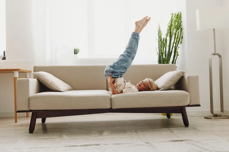 Young woman lies upside down on beige sofa in bright living room, wearing casual jeans and sweater. Cozy home interior with natural light and indoor plants creates relaxed mood.
