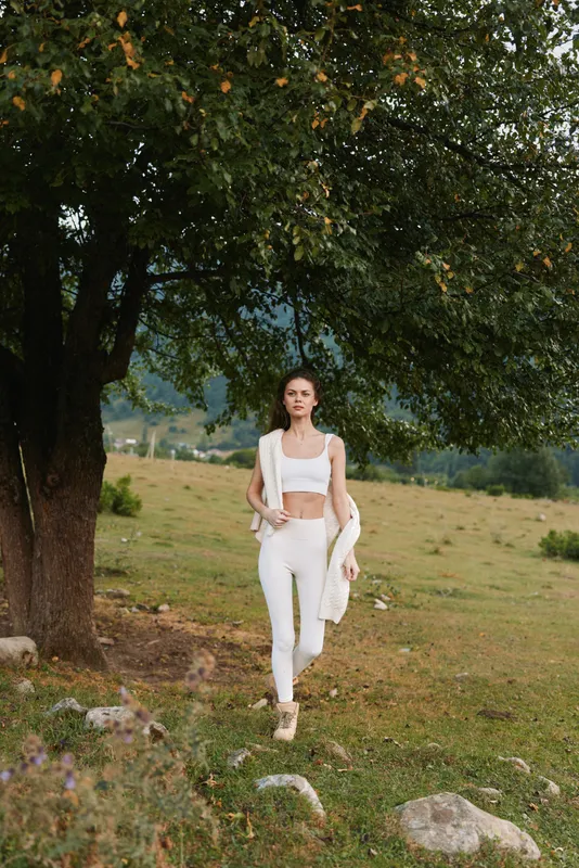 Cheerful woman outdoors, walking through a sunlit meadow in white outfit, relaxed posture, lush greenery, natural colors, candid lifestyle moment warm inviting scene.