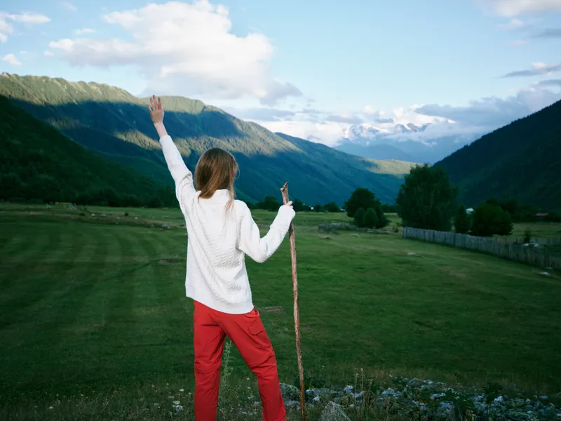 Outdoor scene of a woman standing in a green valley, mountains in the distance, arms raised in triumph, conveying freedom, exploration and joy in vast natural scenery.